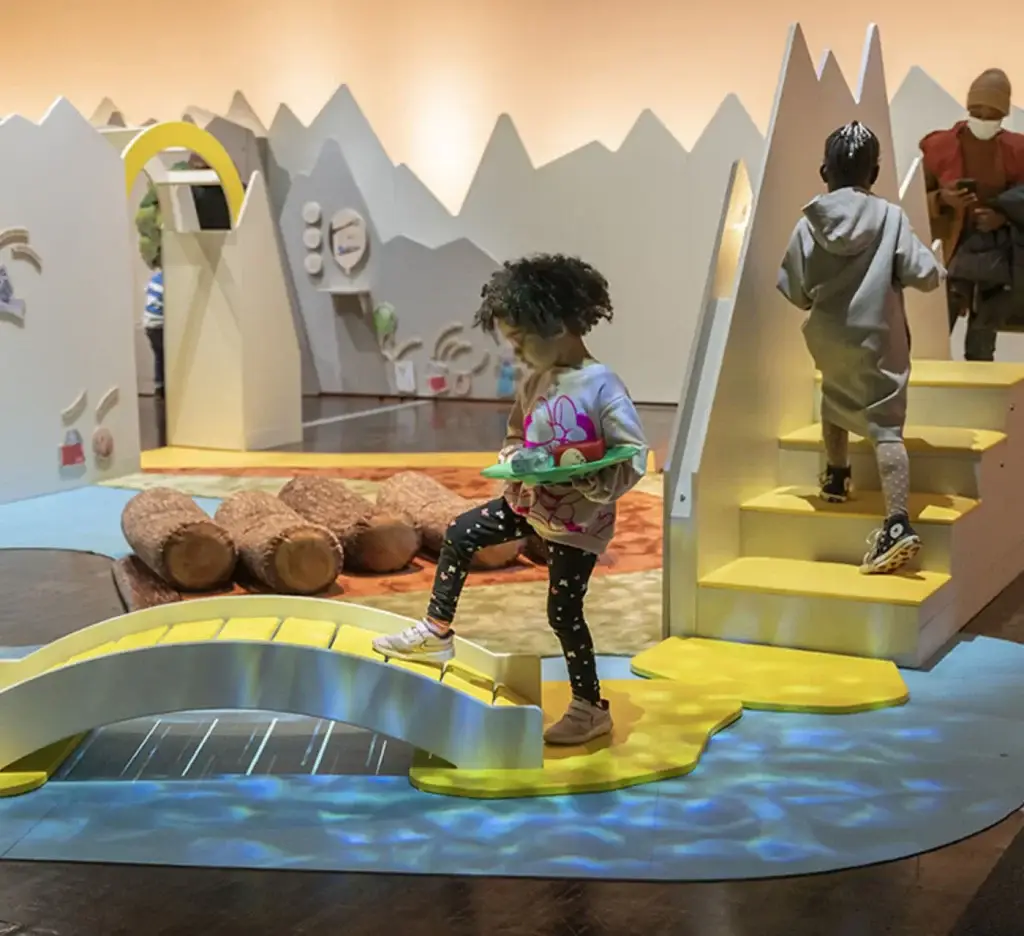 Enfants qui explorent le Nuage des petits au musée des Confluences à Lyon : pont jaune, rivière projetée au sol et petites montagnes