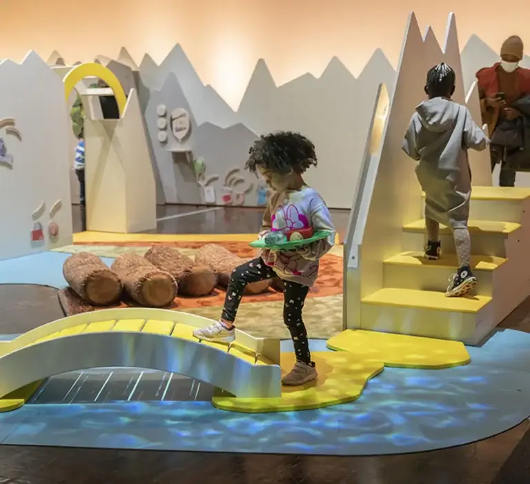 Enfants qui explorent le Nuage des petits au musée des Confluences à Lyon : pont jaune, rivière projetée au sol et petites montagnes